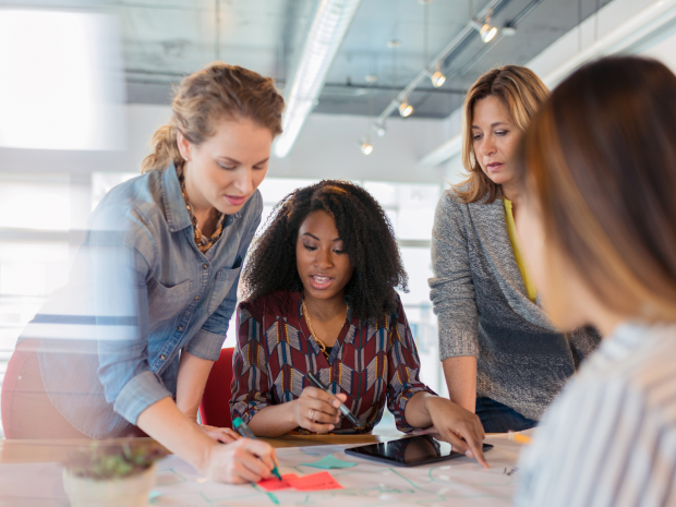 Four people in a conference room having a discussion as one writes on a sticky note.