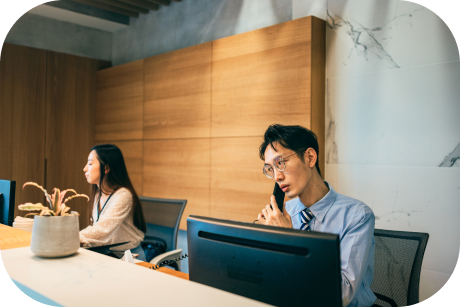 Two people seated in front of desktops one on the phone